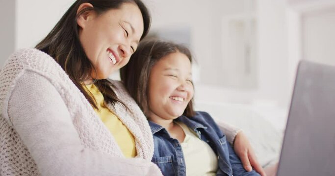 Video of happy asian mother and daughter sitting on sofa with laptop
