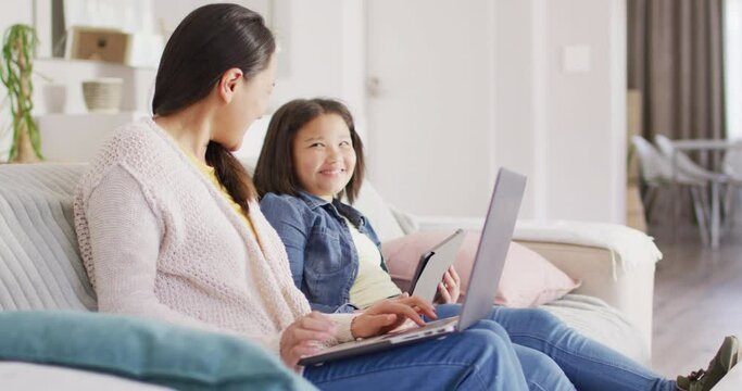 Video of happy asian mother and daughter sitting on sofa with laptop and tablet