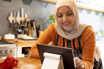 Happy biracial woman wearing hijab, standing in kitchen, using tablet and baking