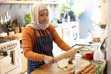 Happy biracial woman wearing hijab, standing in kitchen and baking