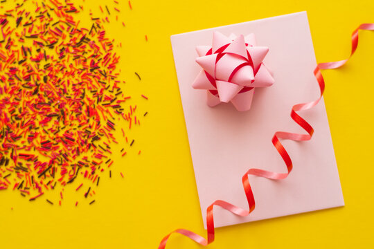 Top view of pink greeting card with gift bow and blurred sprinkles on yellow background.