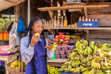 young black African businesswoman in a local market eating apple browsing online using smartphone...