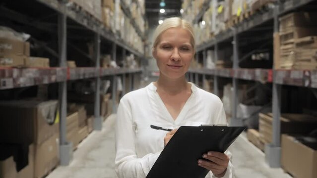 A professional worker, a woman in a white shirt, checks stocks at a retail warehouse. Concept delivery, distribution center. Front view, a woman fills out documents and smiles at the camera.4k footage