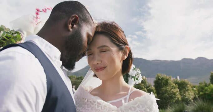 Video of happy diverse bride and groom touching heads together and smiling at outdoor wedding