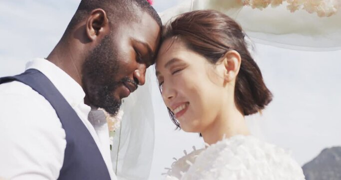 Video of happy diverse bride and groom smiling and touching heads together at outdoor wedding