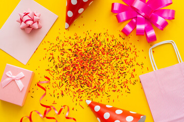 Top view of festive gift near party caps and sprinkles on yellow background.