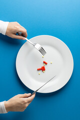 Vertical of hands holding fork and knife over plate with pills, on blue background with copy space
