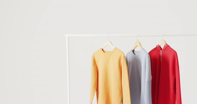 Video of close up of yellow, blue and red sweaters hanging on clothes rail on white background