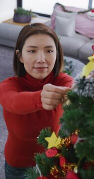 Vertical video of happy asian woman decorating christmas tree at home