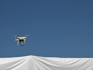 Fototapeta na wymiar white drone in clear blue sky flying over a white awning, launching a copter into the airspace over a white rooftop, conceptual image of a four propeller drone flying over a white canopy