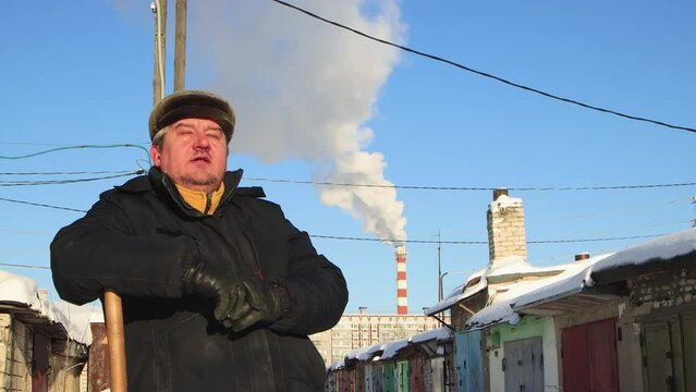 A man in a fur hat rests leaning on a wooden snow shovel. Large snowdrifts around after a snowstorm, sunny day. City life, heavy snowfall. UHD 4K.