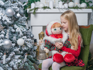 A little blonde girl is sitting on a chair in a Christmas interior. The girl is holding a doll in her hands. Magical Christmas time. Horizontal photo.