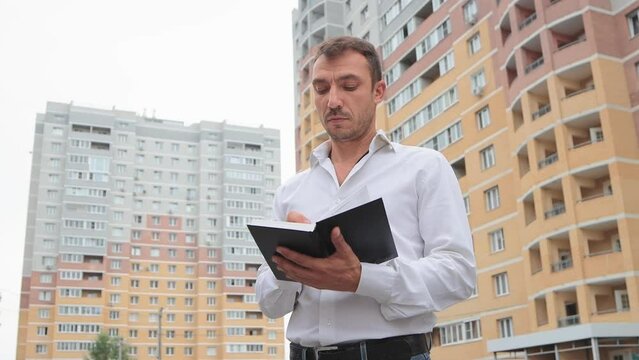 Focused businessman in a white shirt makes notes in a notebook. A young man in the city center against the backdrop of high-rise buildings. Business concept remotely, online. 4K UHD.
