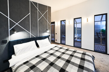 Contemporary bedroom with numerous glass doors leading to terrace at dusk. Side view of illuminated big bed with plaid blanket in front of gray wall in sleeping area indoors. Concept of interior.