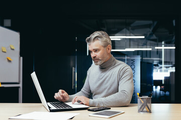 Serious and pensive mature investor working inside modern office, senior gray-haired businessman using laptop at work, male boss at work sitting at desk.