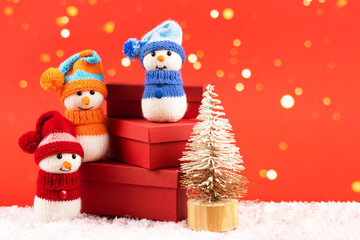 Knitted snowmen in a red hat and sweater stands on three red gift boxes on a red background with golden Christmas lights. Merry Christmas and New Year 2023 greeting card