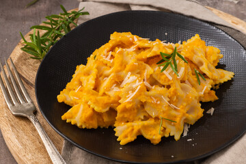 Farfalle pasta with pumpkin sauce and parmesan cheese decorated with rosemary on a black ceramic plate. Creamy cheesy vegan pumpkin pasta. Kitchen counter top.
