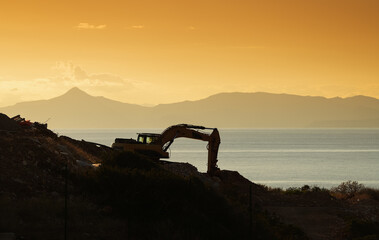 Fototapeta na wymiar Silhouette of an excavator digging on the sea shore against sunset sky. Construction industry heavy machines.