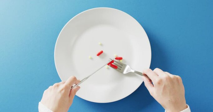 Video of hands holding knife and fork over dinner plate of various pills on blue background