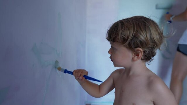 Child paints wall with paintbrush. Cut small boy helping mother with house renovation. Kid and parent painting home wall
