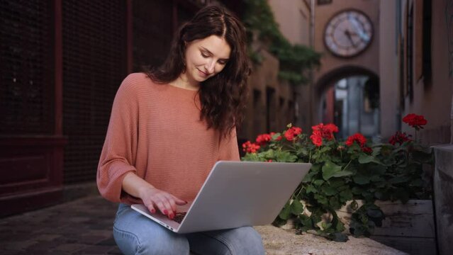 using laptop, hands typing, Woman worker. An outdoor student is studying, a freelancer is sitting on decorative stone in a city street, and acute, cheerful, smiling, romantic blogger is working