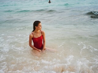 Sexy woman in a red swimsuit with an athletic body stands in the water in the ocean and poses looking out at the horizon