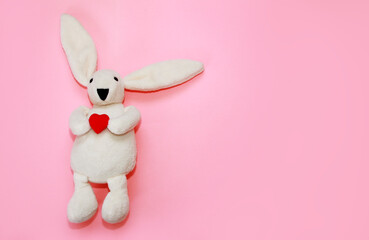 children's toy soft fluffy white rabbit holding a red heart on a pink background