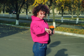 Obraz premium Smiling young woman with curly hair in magenta hoodie holds cup with coffee outdoors