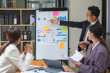 Team business three smiling and happy Asian men and women team using tablets and laptops brainstorm and plot statistics on a corporate chart board inside the company. Colleagues concept.