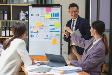 Team business three smiling and happy Asian men and women team using tablets and laptops brainstorm and plot statistics on a corporate chart board inside the company. Colleagues concept.