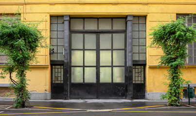 Fototapeta na wymiar Frontal view of a yellow factory facade with a nostalgic black gate with glass panes in Budapest Hungary. In the foreground is a wet road and two street lamps overgrown with ivy