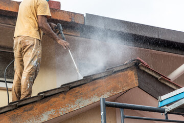 Worker using high pressure water jet spray gun to wash and clean dirt from rooftop tiles in...