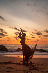 Silhouette slim woman dancing arms raised on sandy beach at sea background, posing. Happy lady wearing black dress dance on tropical ocean at sunset. Travel vacation concept. Copy text space