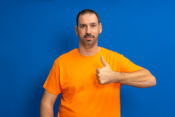 Bearded handsome hispanic man wearing orange casual t-shirt standing over isolated blue background doing happy thumbs up gesture with hand. Approval expression looking at camera showing success