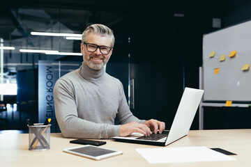 Fototapeta na wymiar Portrait of successful senior businessman, gray haired man in glasses smiling and looking at camera, mature investor boss working inside modern office building using laptop at work.