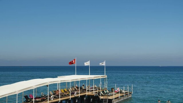 Pier with white awning and sun loungers goes into the Mediterranean Sea. Against the background of the mountain. The flag of Turkey and the hotel are developing in the wind. People swimming in sea.