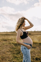 Blonde pregnant woman wearing a top and jeans, standing in an open field, looking away.