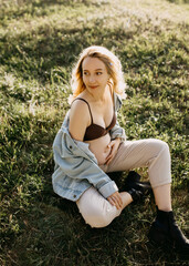 Fototapeta premium Blonde pregnant woman wearing a denim jacket, sitting in an open field, holding hand on belly, smiling.