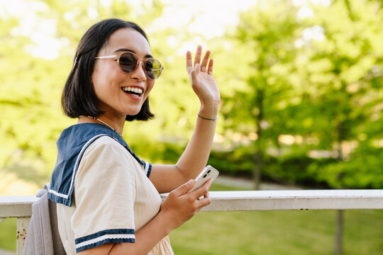 Young beautiful asian girl using mobile phone and waving hand while standing in park