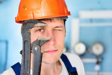 Professional plumber in helmet looks directly into camera through large adjustable wrench. Large portrait of real worker 30 years old..