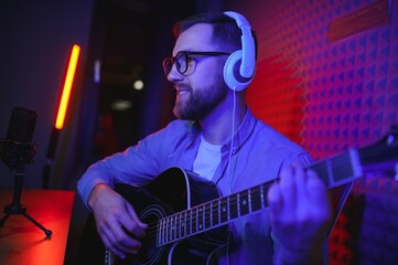 Obraz premium A young stylish guy with a beard wearing casual or music teacher playing guitar in front of smartphone camera. Online guitar training or vocal lesson