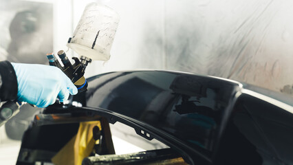 Close-up indoor shot of the process of fender varnishing with black automobile painting. Hand in blue rubber glove holding paint spray gun. High quality photo