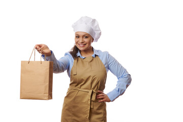 Adorable woman waitress or chef confectioner, baker in blue casual shirt and beige apron, smiling and holding out a disposable eco paper bag with fresh delivered food, isolated over white background