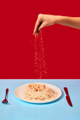 Female hand salting plate with pearl necklaces symbolizing noodles, pasta on blue tablecloth over red background. Food pop art photography.