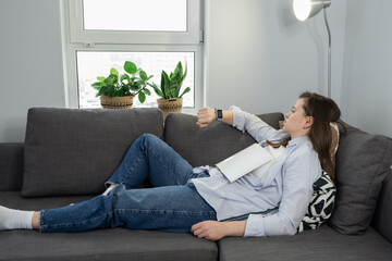 Woman on sofa at home with book on her chest, taking a break, looking at her smart watch. Fatigue and sleep disorder concept