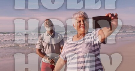 Animation of christmas greetings text over senior biracial couple taking selfies on beach - Powered by Adobe