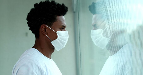 Black man in lockdown wearing covid-19 mask looking out apartment window, african person during quarantine