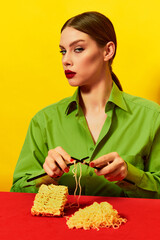 Woman with emotionless face knitting instant noodles on blue table on vivid red tablecloth over yellow background. Food pop art photography.