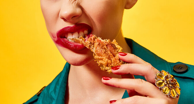 Naklejki Cropped image of woman in green coat with red lipstick smudge, eating fried chicken, nuggets over yellow background. Spicy taste. Food pop art photography.