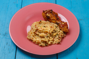 Couscous in sauce and spices with grilled chicken drumstick on a plate on a blue table.